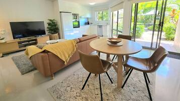 A modern living room with a sofa, a TV, a dining table with chairs, and a kitchen in the background.