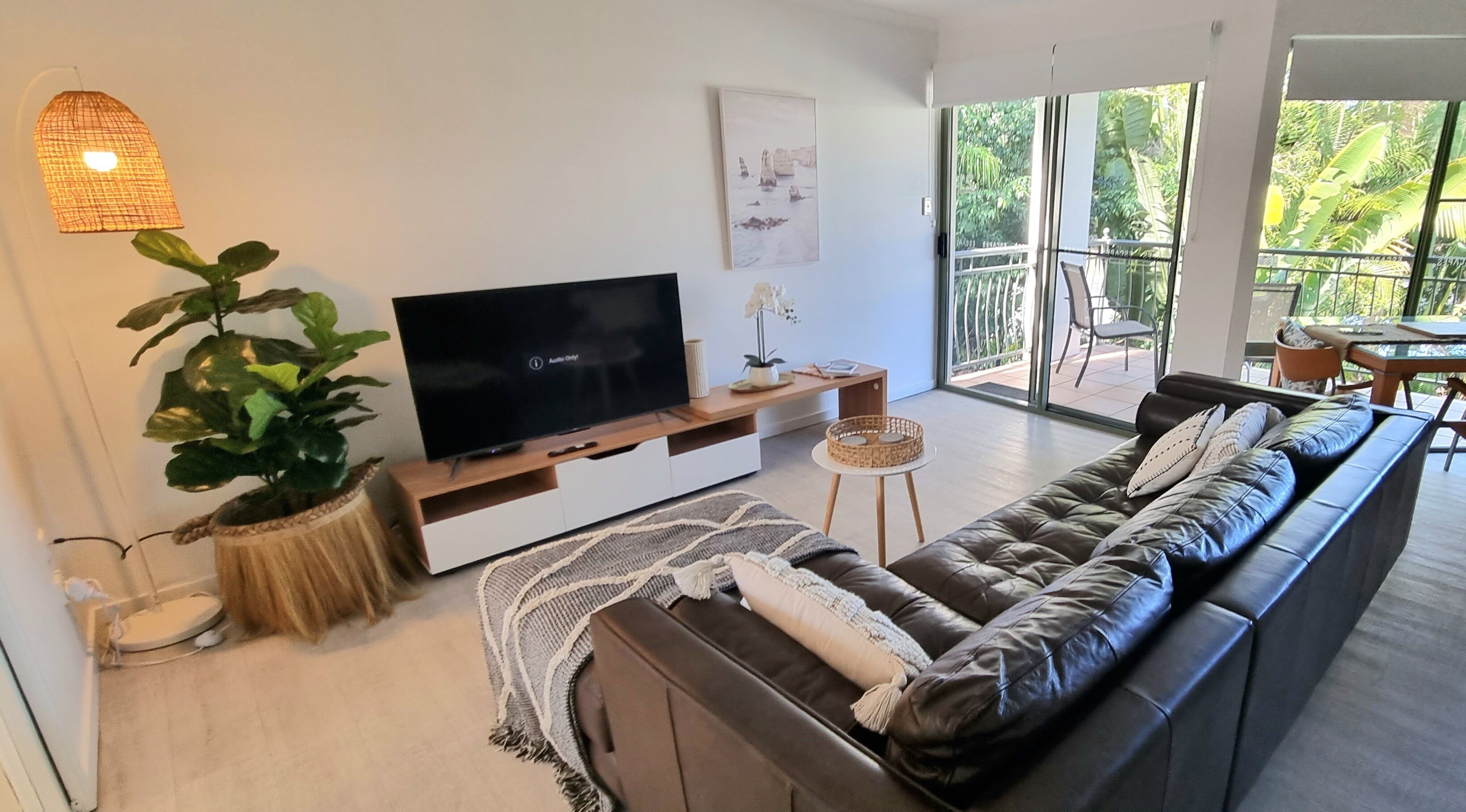 A modern living room with a large flat-screen TV, a comfortable sofa, a potted plant, and a balcony with a view of greenery.