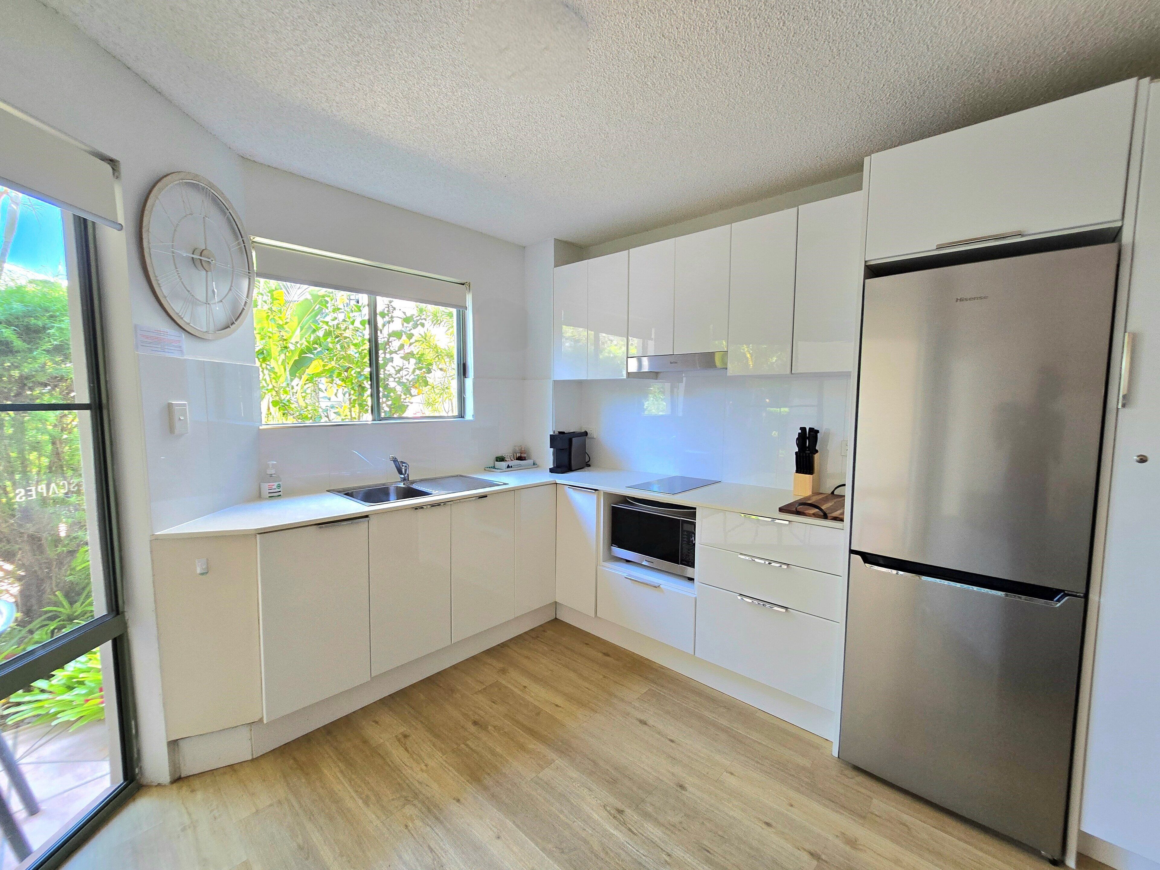 A modern kitchen with white cabinets, a stainless steel refrigerator, a built-in oven, and a window with a view of greenery.