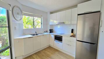 A modern kitchen with white cabinets, a stainless steel refrigerator, a built-in oven, and a window with a view of greenery.