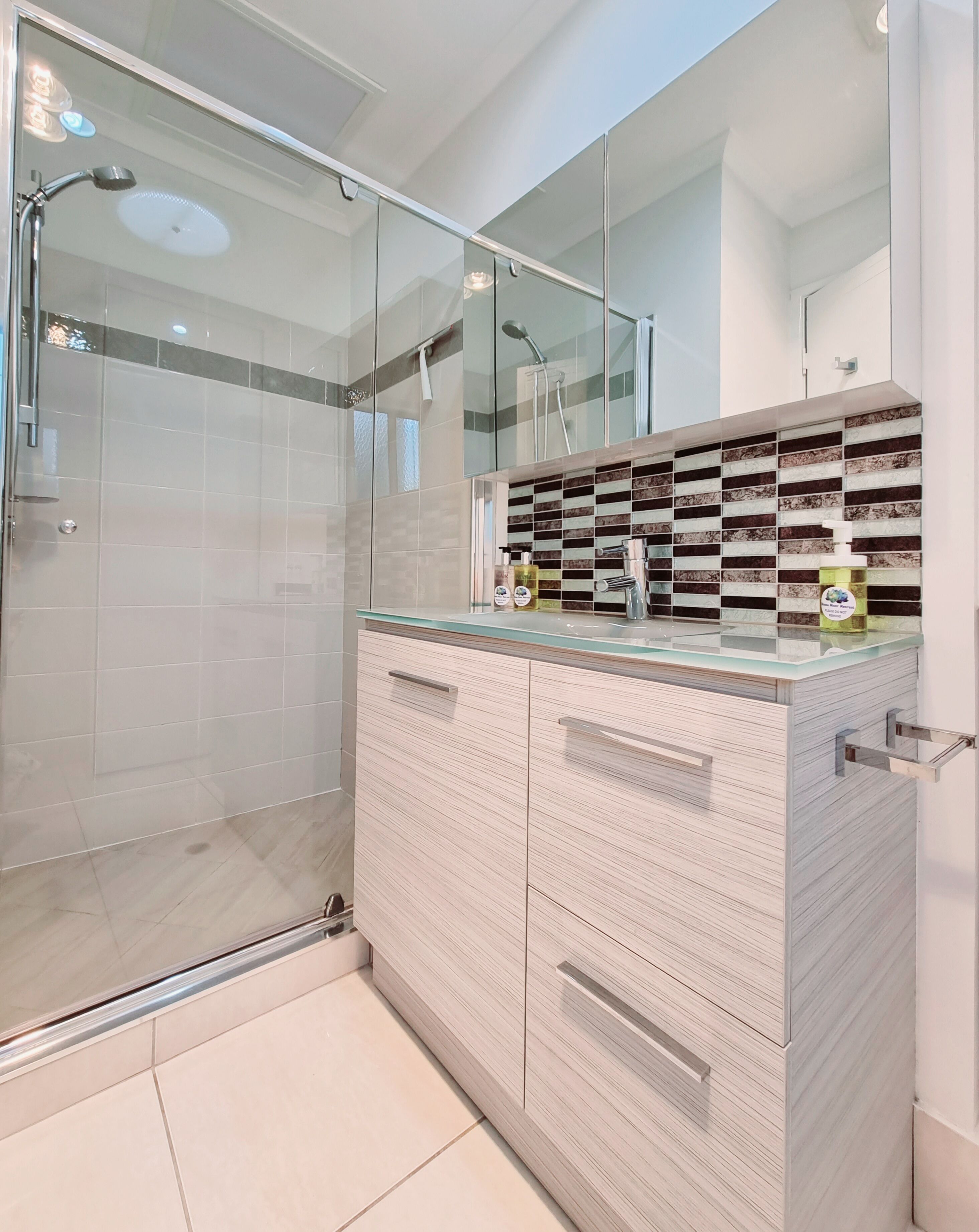 Une salle de bain moderne dotée d’une douche à parois de verre, d’un meuble-lavabo avec un plateau en verre et d’un mur recouvert de carreaux.