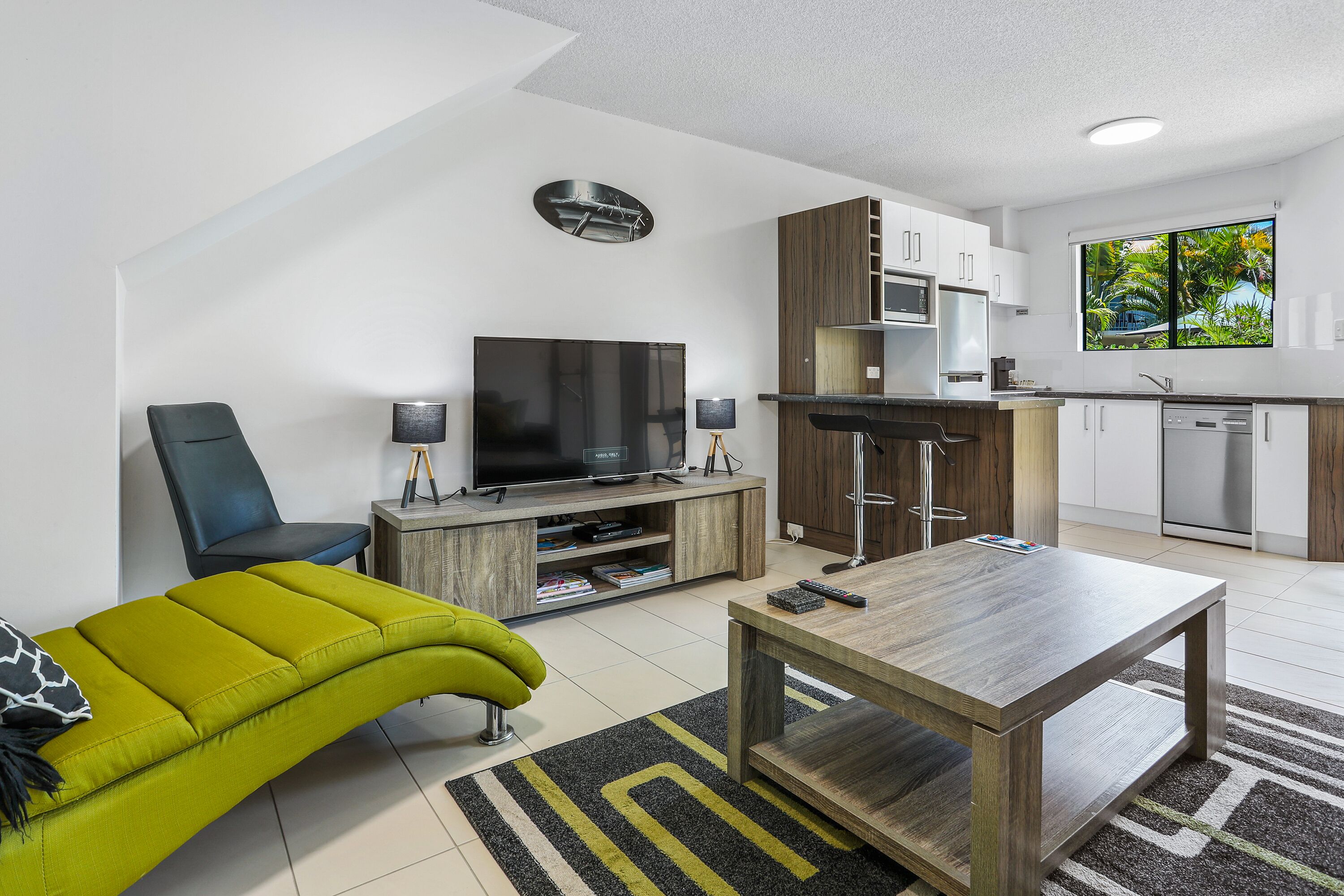 A modern living room with a flat-screen TV, a green sofa, a wooden coffee table, and a kitchen area with white cabinets and a window with a tropical view.