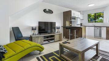 A modern living room with a flat-screen TV, a green sofa, a wooden coffee table, and a kitchen area with white cabinets and a window with a tropical view.
