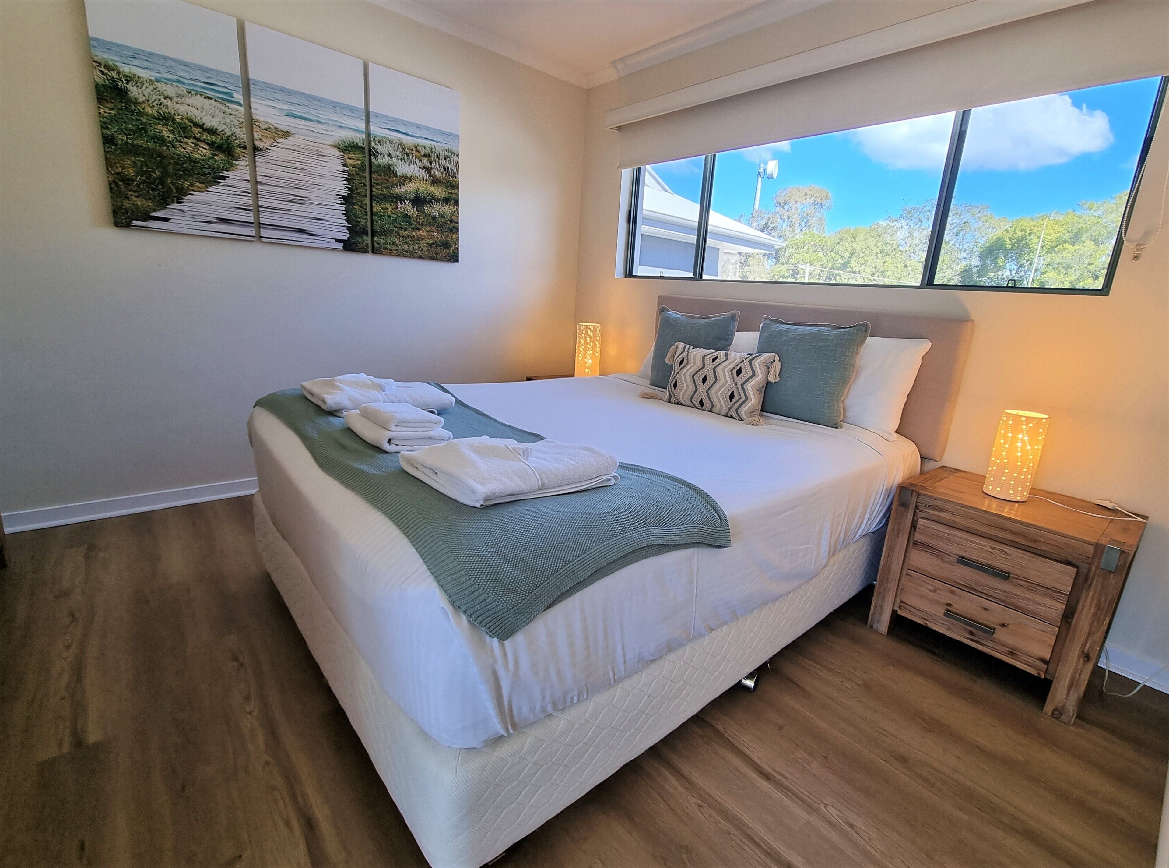 A neatly made bed with white linens and a green throw, a bedside table with a lamp, and a large window with a view of trees and a clear sky.