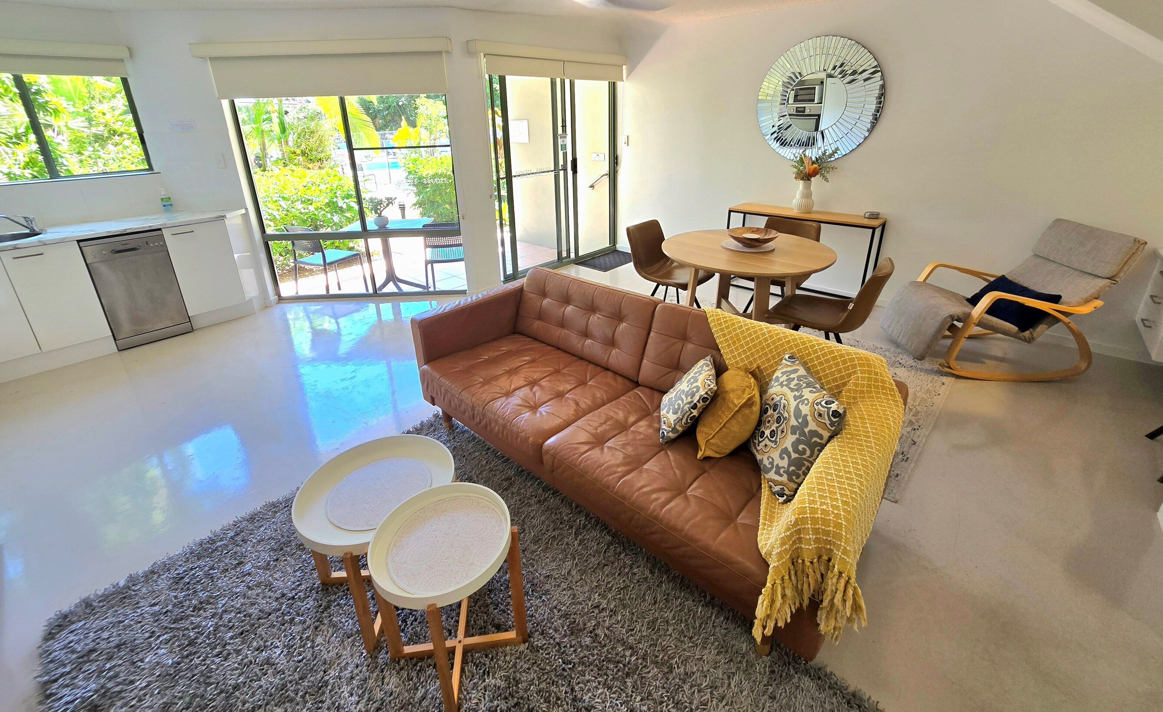 A modern living room with a brown leather sofa, two small side tables, a rocking chair, and a dining area with a round table and chairs.