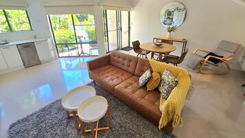A modern living room with a brown leather sofa, two small side tables, a rocking chair, and a dining area with a round table and chairs.