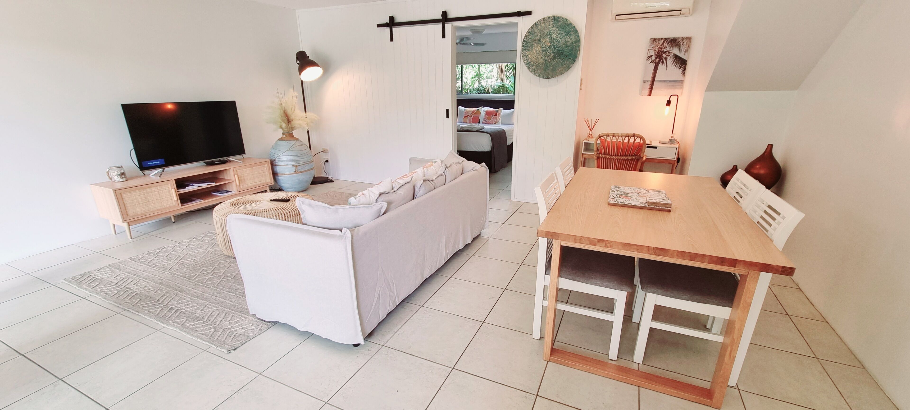 A modern living room with a flat-screen TV, a sofa, a dining table, and a kitchen area visible through an open door.