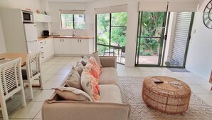 Una cocina moderna con armarios blancos, una zona de comedor con una mesa y sillas de madera, un sofá con cojines de colores, un reposapiés de mimbre y un televisor sobre un mueble de madera.