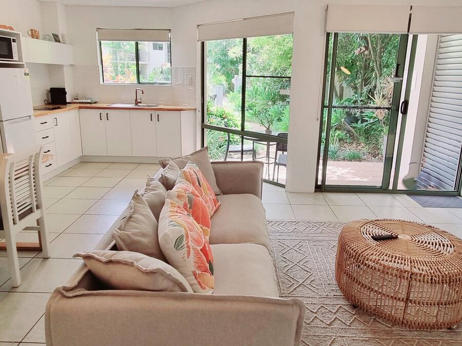 A modern kitchen with white cabinets, a dining area with a wooden table and chairs, a sofa with colorful cushions, a wicker ottoman, and a TV on a wooden cabinet.
