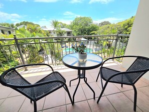 Un balcon avec deux fauteuils en osier noir, une petite table ronde ornée d’un pot de fleurs, et une vue sur une piscine et la verdure environnante.