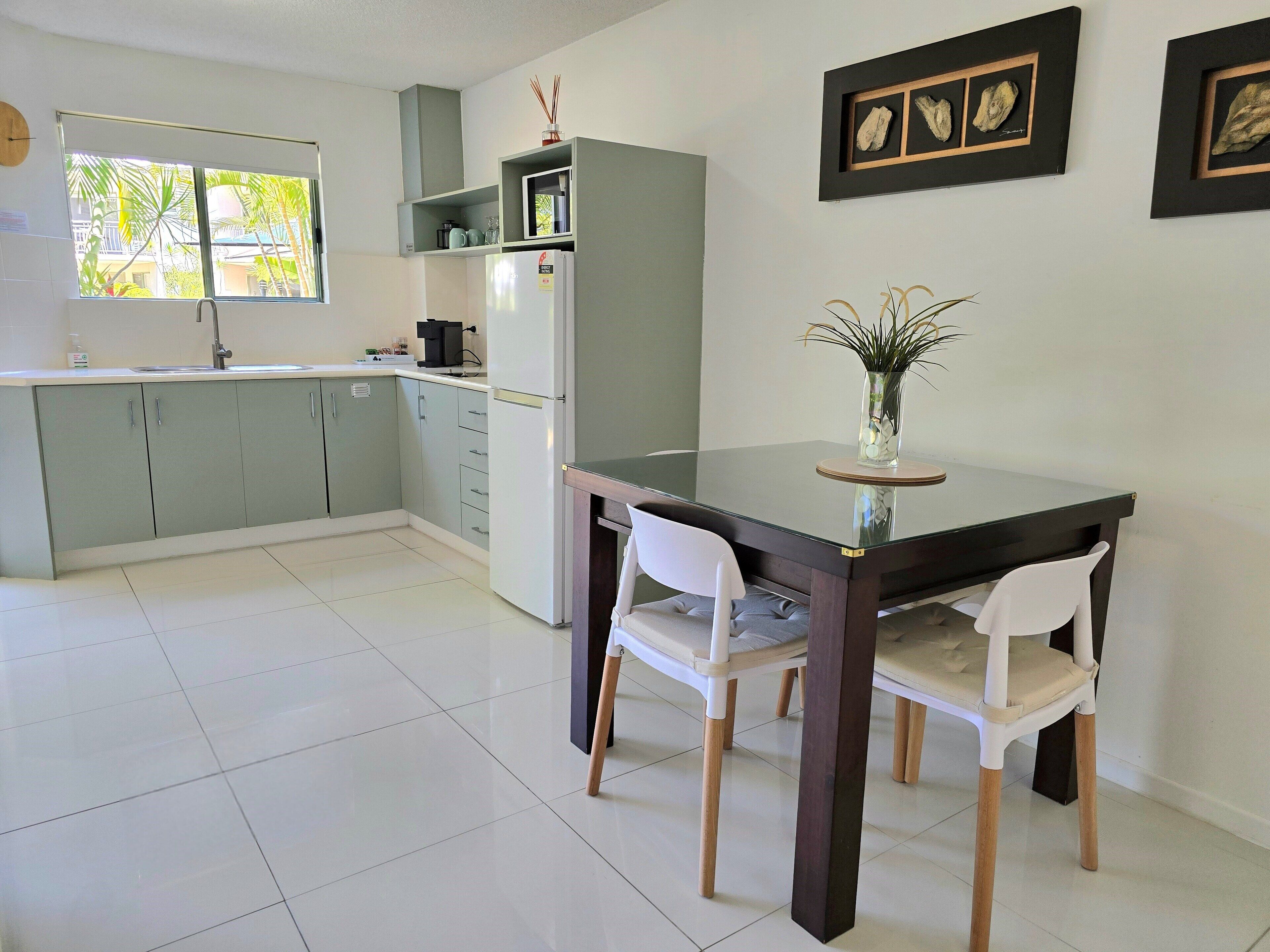 Una cocina moderna con armarios grises, un refrigerador blanco y una mesa de comedor con dos sillas blancas. Hay cuadros en la pared y una ventana con vistas a palmeras.