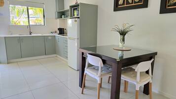 A modern kitchen with grey cabinets, a white refrigerator, and a dining table with two white chairs. There are framed pictures on the wall and a window with a view of palm trees.