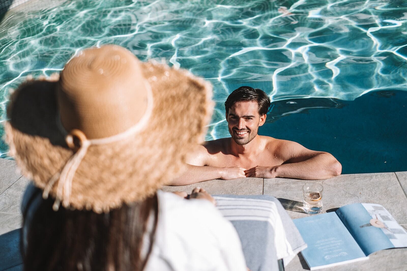 A person in a swimming pool with a book and a glass of drink on the edge.