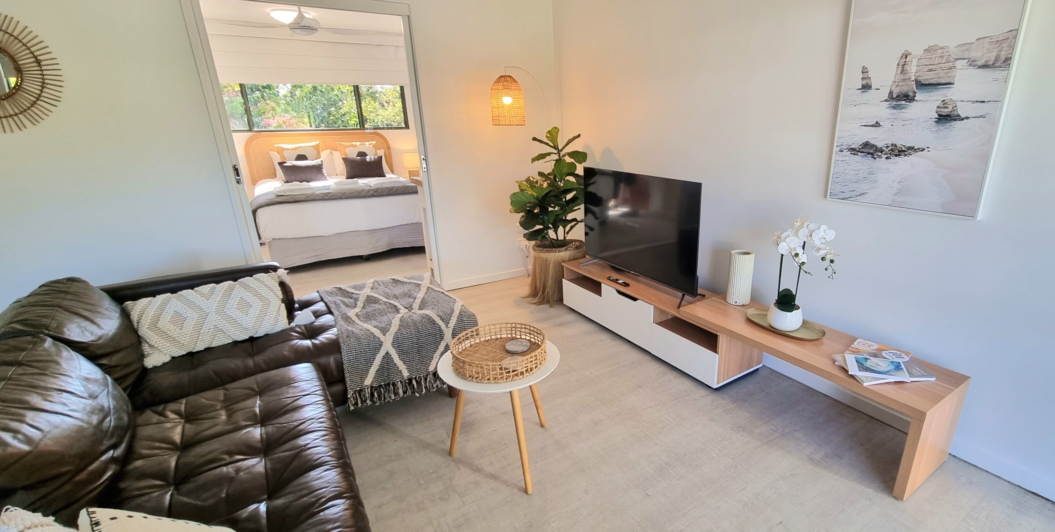 A modern living room with a leather sofa, a TV on a wooden stand, a small round table, and a bed with a view to the outdoors.