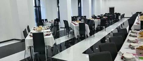 A large conference room with long tables set for a meal, equipped with chairs and tables, and a modern ceiling with recessed lighting.