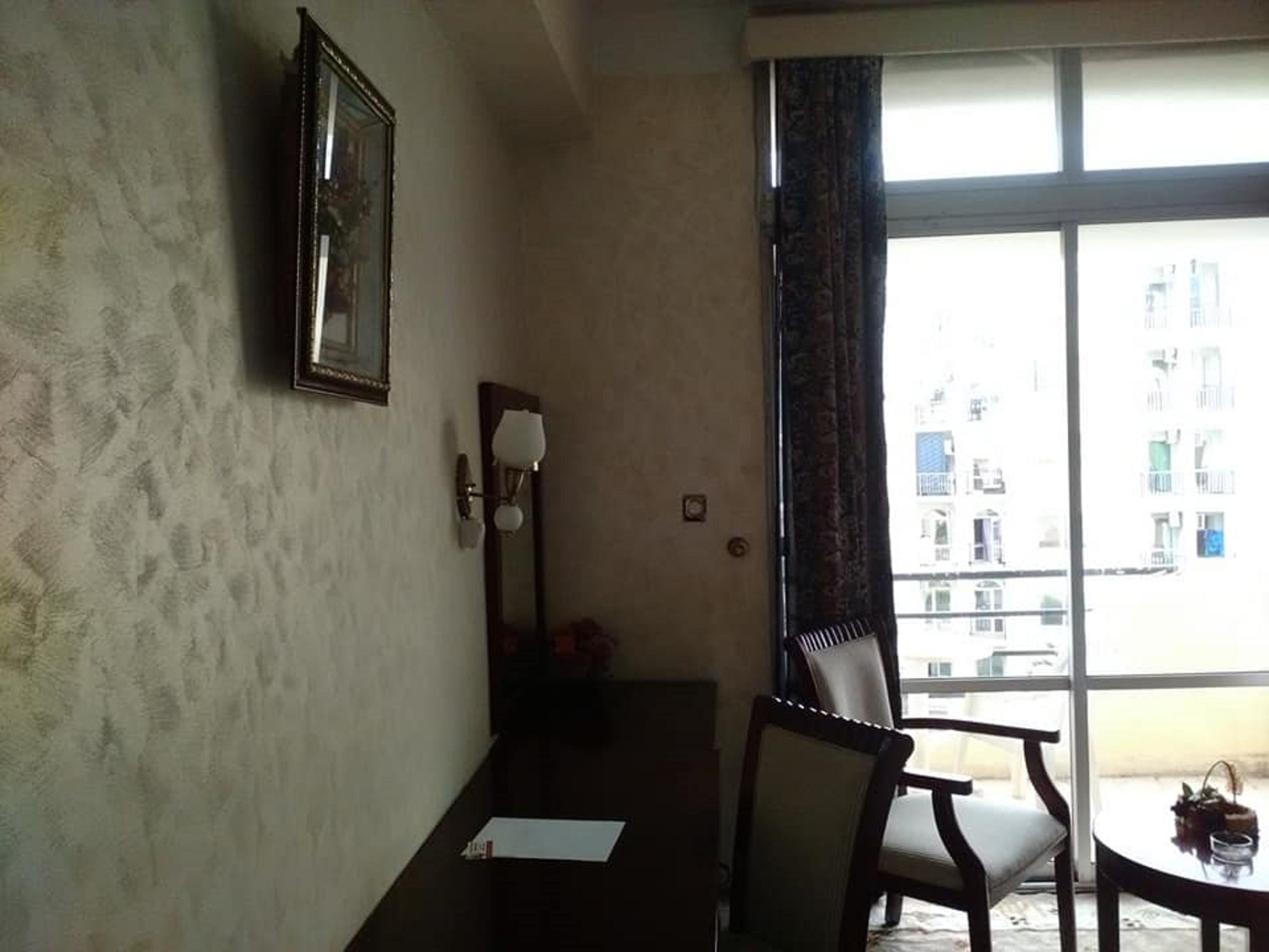 A hotel room with a large window, a framed picture on the wall, a desk with a chair, and a small table with a vase.