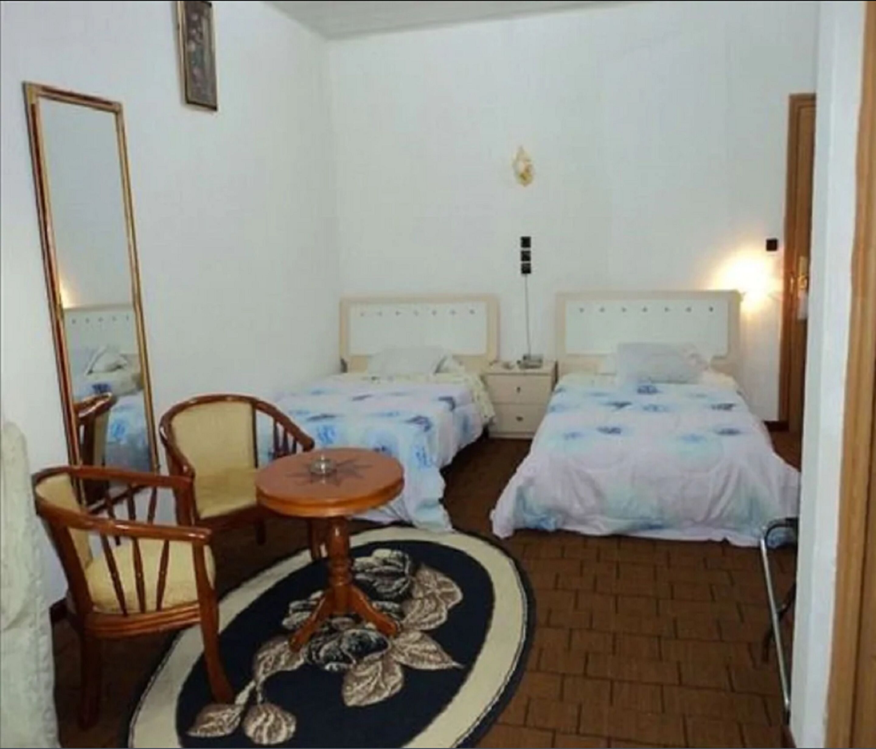 Habitación de hotel con dos camas individuales, una mesa redonda con camino de mesa floral, una silla de madera y un espejo grande.