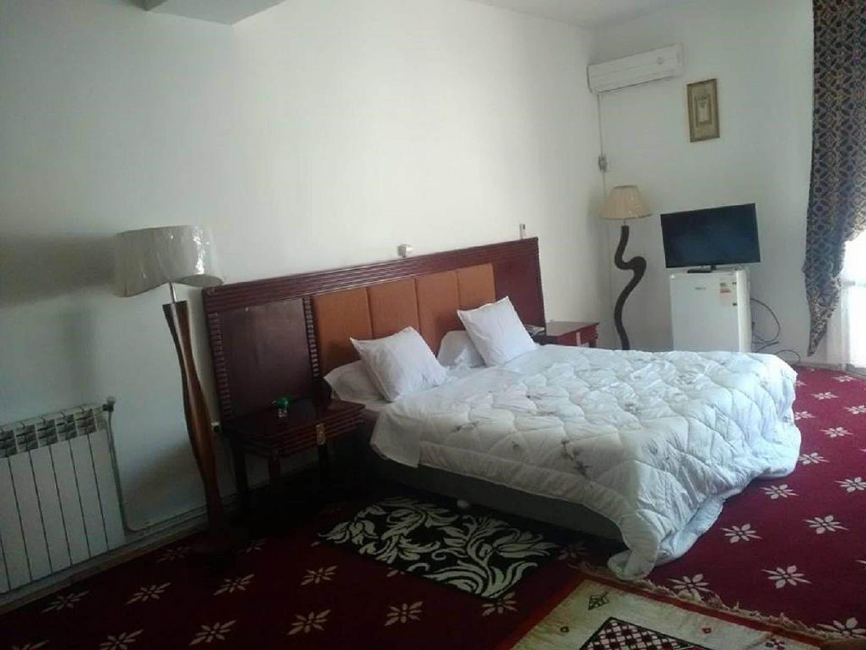 Habitación de hotel con cama de madera, ropa de cama blanca, alfombra roja, televisor y radiador.