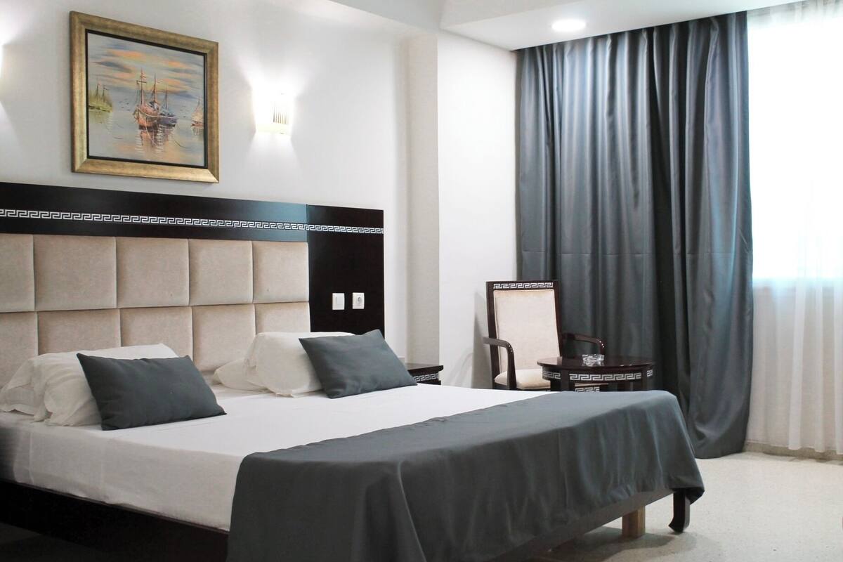 Habitación de hotel con una cama grande, cabecera con diseño geométrico, un cuadro en la pared, una mesita de noche y una ventana con cortinas.