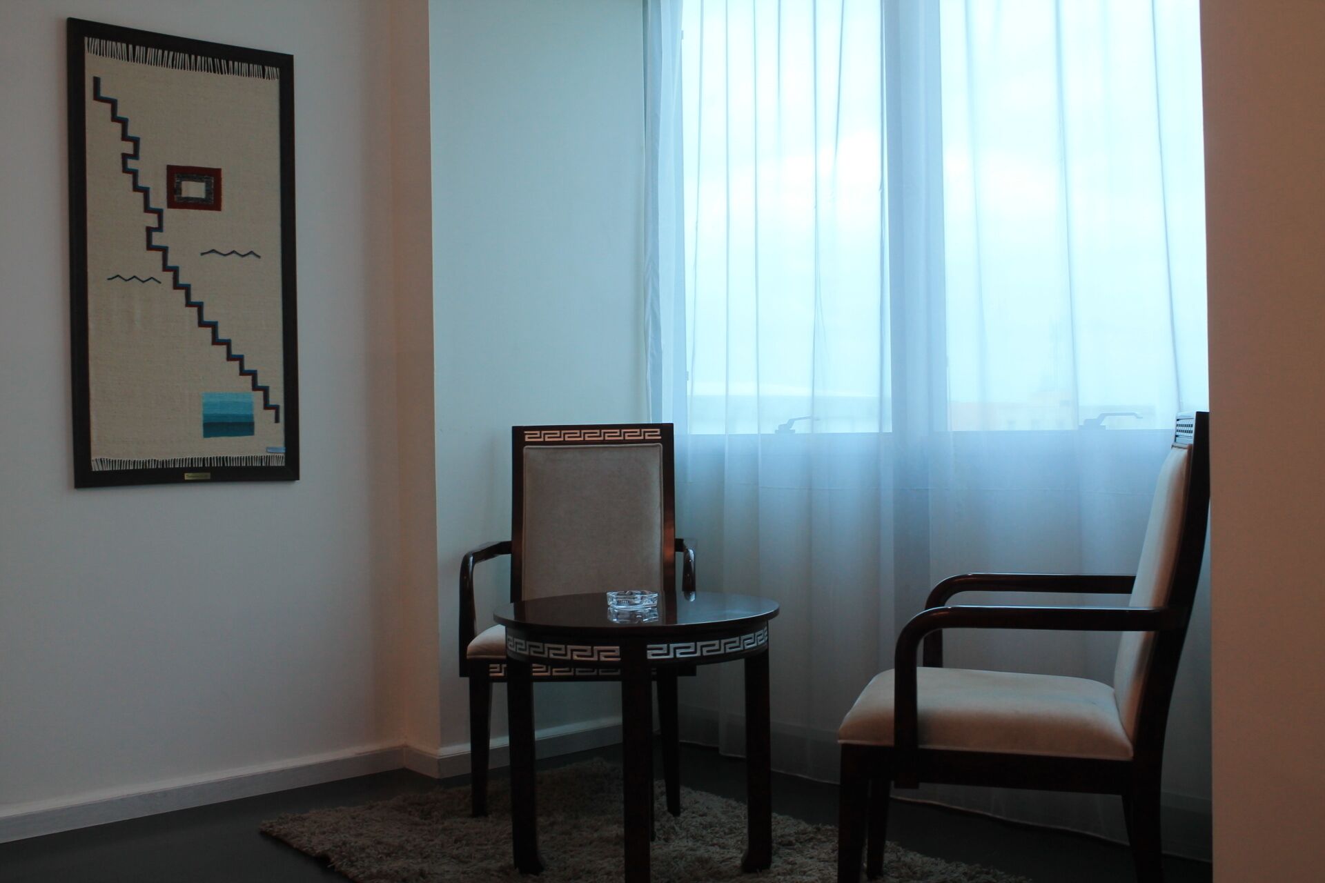 A room with a window, a chair, a table, and a framed artwork on the wall.