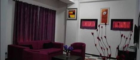 A living room with a red sofa set, a coffee table, and a patterned rug. There are two framed pictures on the wall and a vase with red flowers on a side table.
