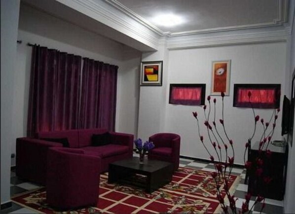 A living room with a red sofa set, a coffee table, and a patterned rug. There are two framed pictures on the wall and a vase with red flowers on a side table. - Majestic Annaba (Annaba)