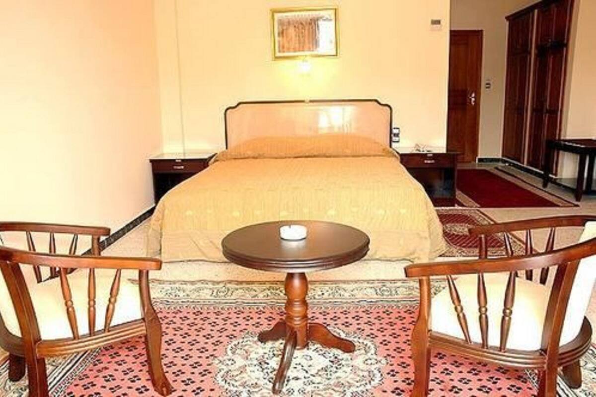 A hotel room with a bed, two wooden chairs, a small round table, and a patterned rug.