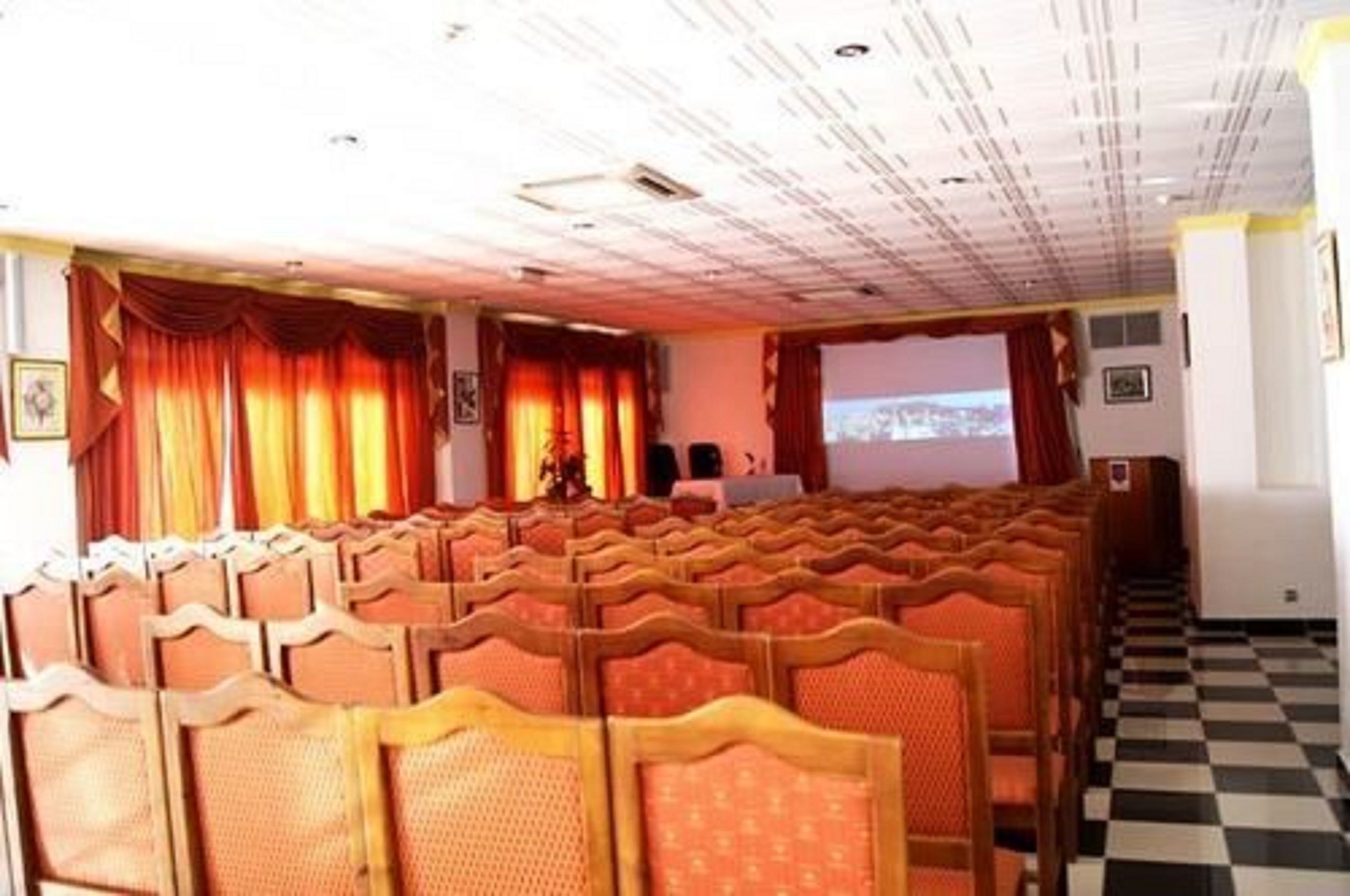 A conference room with rows of wooden chairs, a projector screen, and a checkered floor.