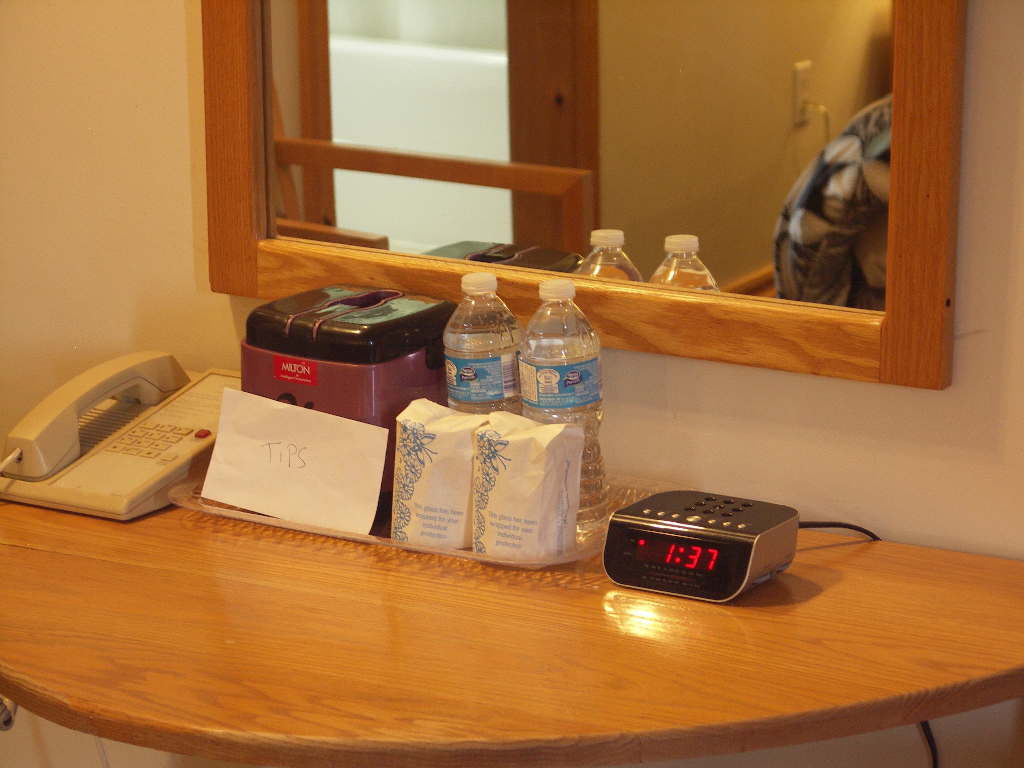 A hotel room counter with a telephone, a minibar, bottled water, a clock displaying 1:37, and a note labeled "TPS".