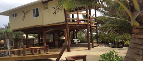A beachside building with a wooden deck, a covered seating area, and a palm tree.