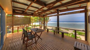 A wooden deck with a table and chairs, overlooking a beach with palm trees and the ocean. - Tropical Sands (Rarotonga)
