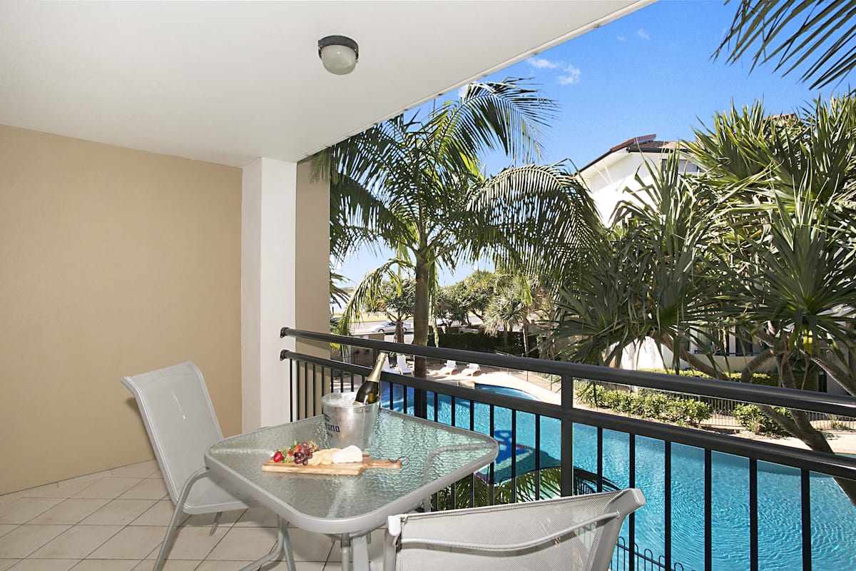 Ein Balkon mit Glastisch, zwei Stühlen, einer Champagnerflasche und Blick auf einen Swimmingpool und Palmen.