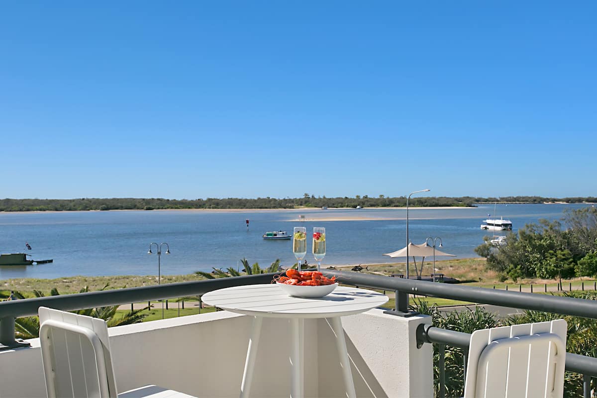 Ein Balkon mit Tisch, Stühlen und einer Schüssel Essen. Im Hintergrund ist Wasser, Boote und eine entfernte Küste zu sehen.