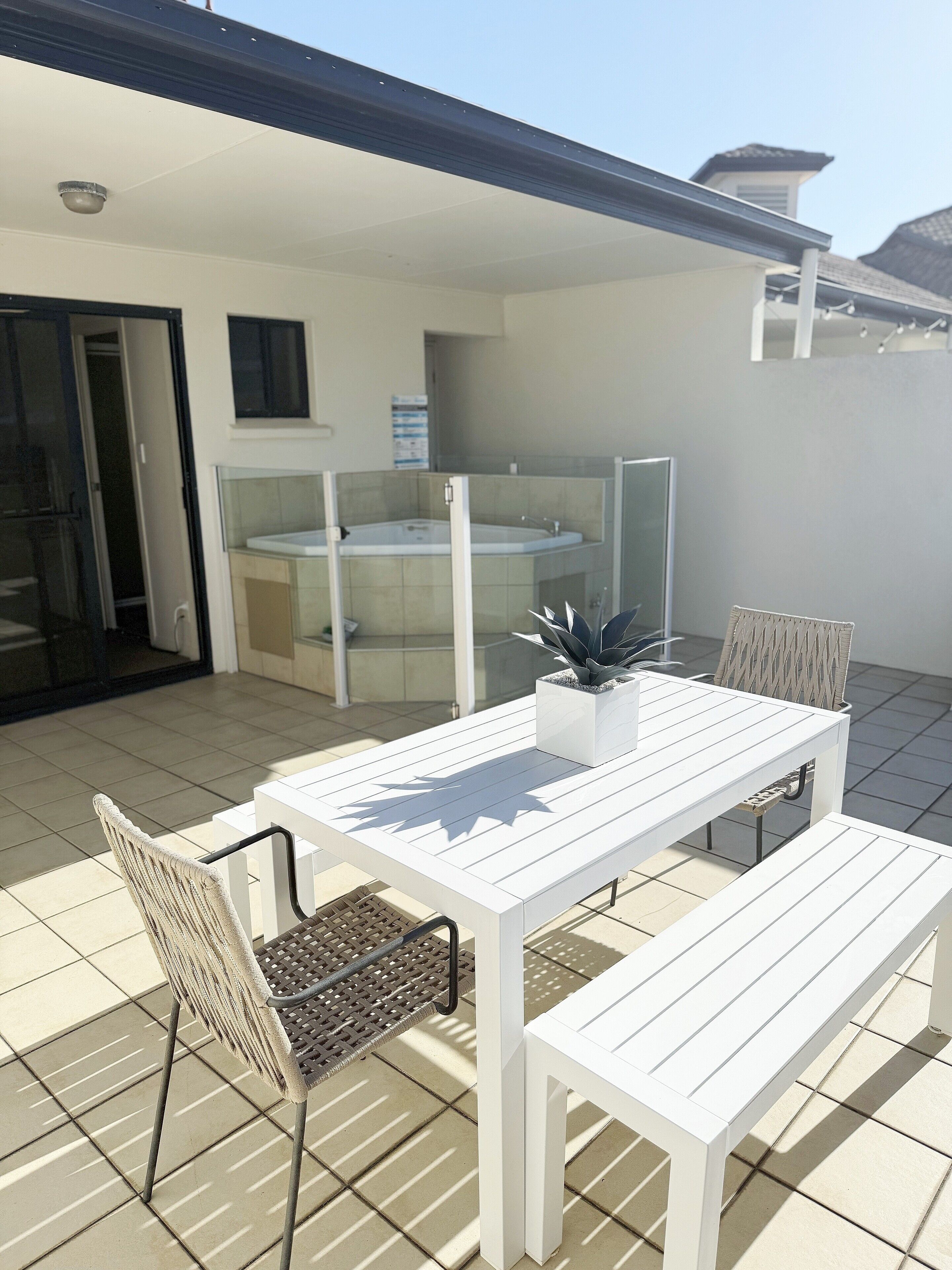 Ein weißer Tisch und Stühle auf einer gefliesten Terrasse mit einem gläsernen Whirlpool und einer Tür, die zu einem Raum führt.