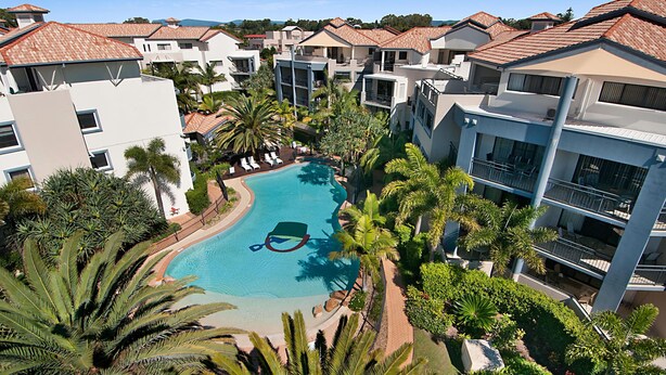 Un resort con piscina rodeada de palmeras y edificios modernos con techos de tejas rojas.