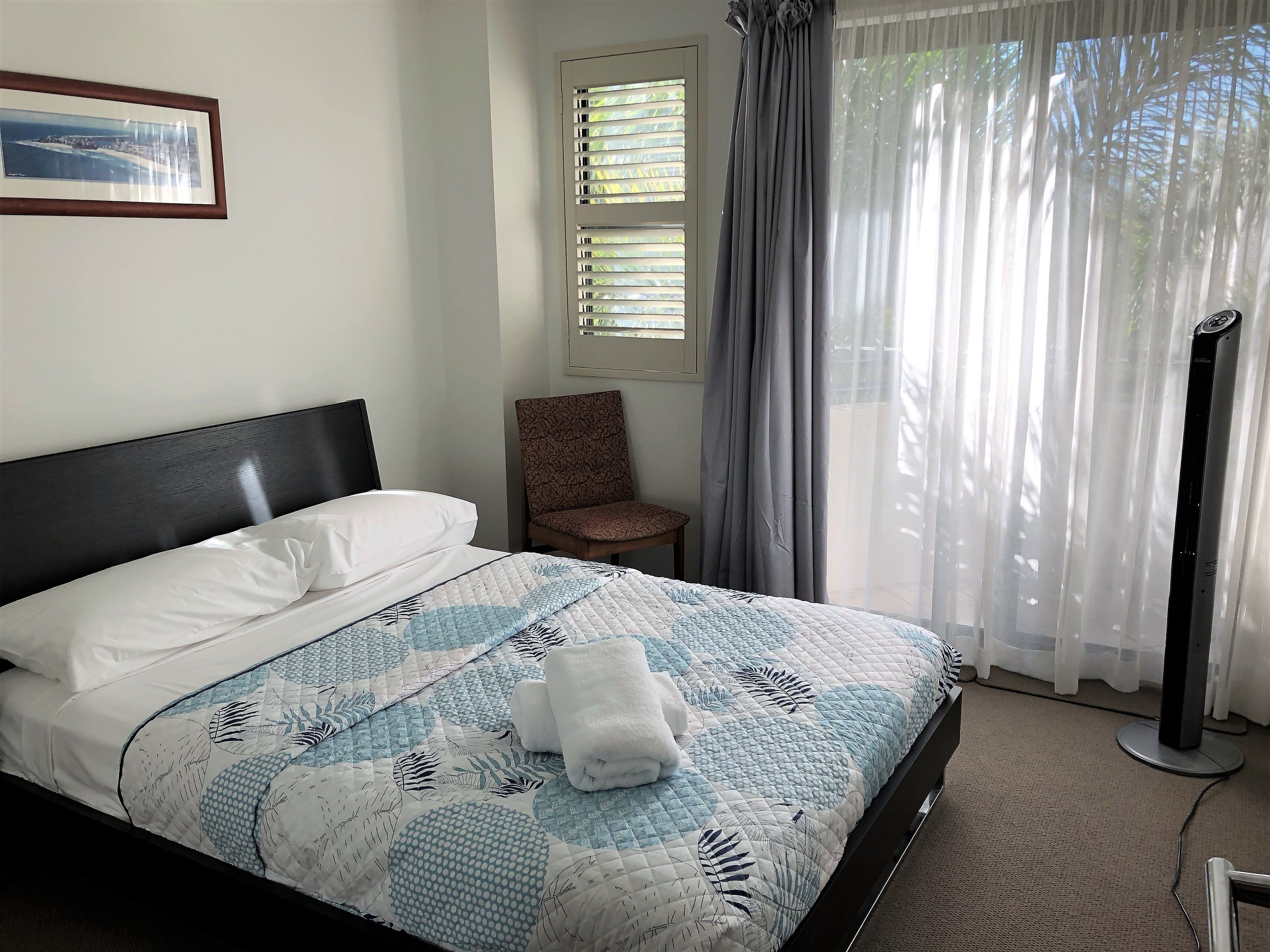 A neatly made bed with a quilt, a chair, a picture on the wall, and a window with curtains.