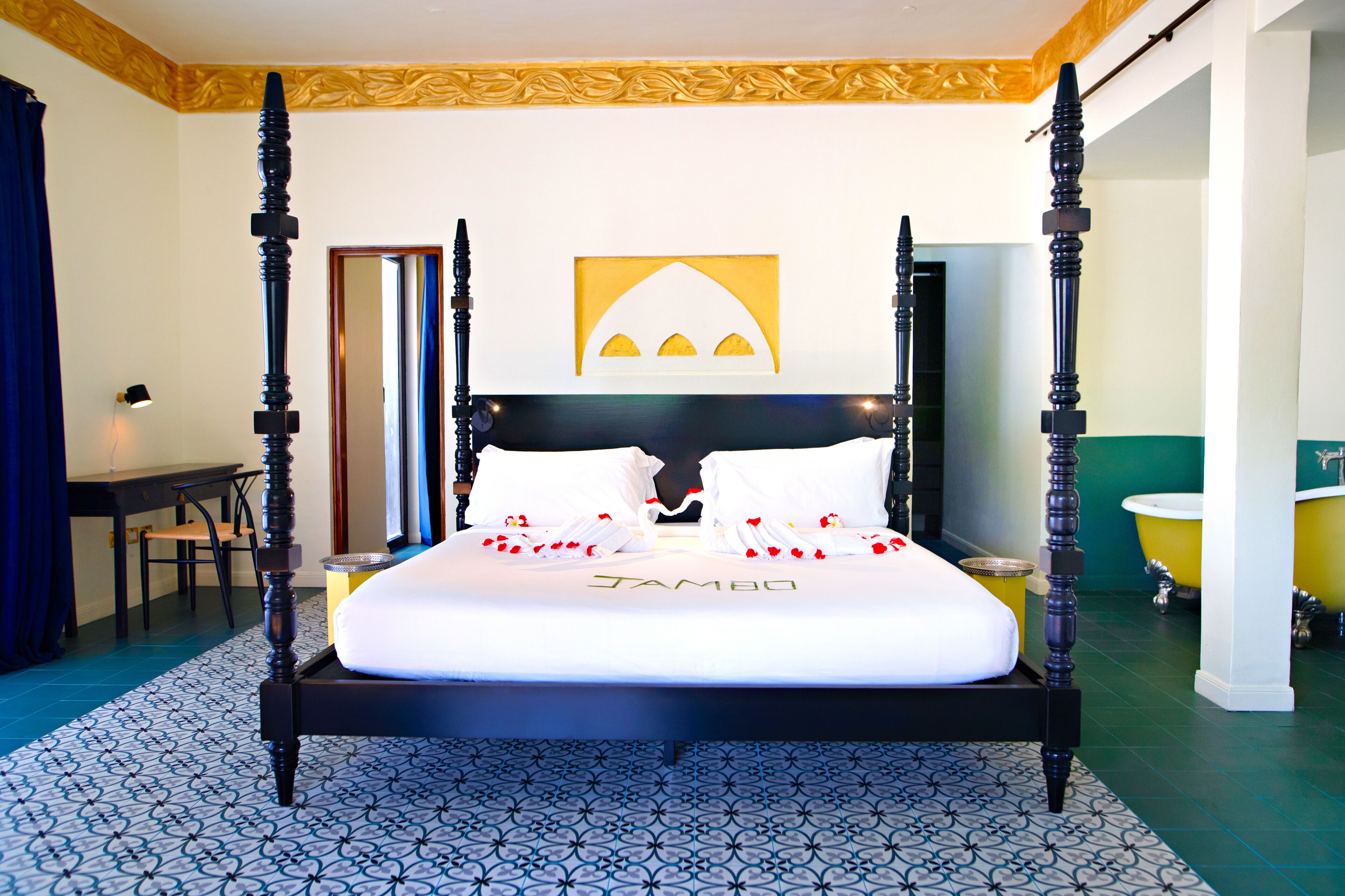 Ein Bett mit Himmel, mit weißer Bettdecke, das Schild "TAMBO" darauf, ein Spiegel über dem Bett, ein Schreibtisch mit Stuhl und eine gelbe Badewanne im Badezimmer.