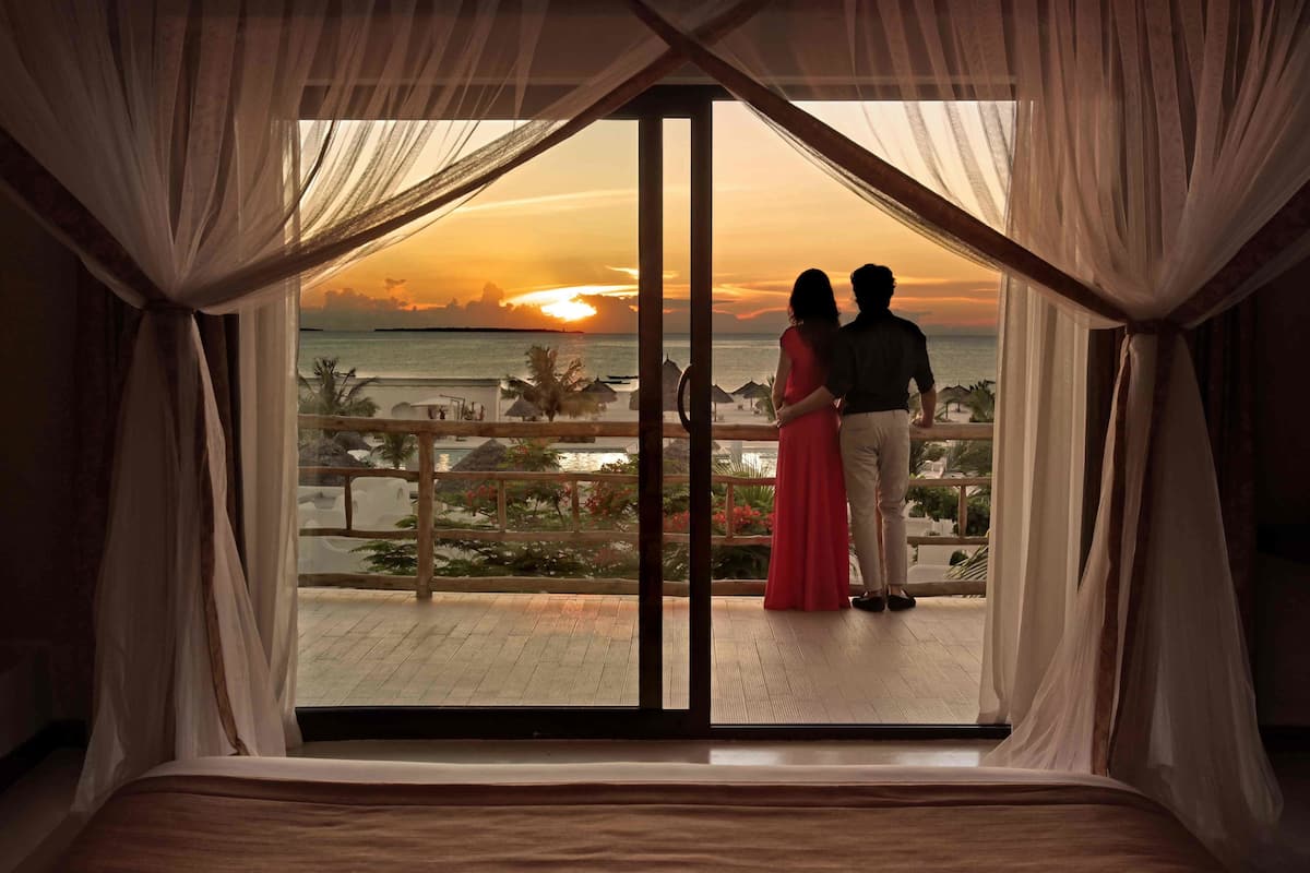 A couple standing by a balcony with a view of the sunset over the ocean.