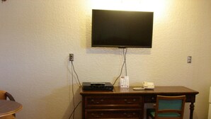 A room with a flat-screen TV mounted on the wall, a wooden desk with a chair, and a small table with a chair.