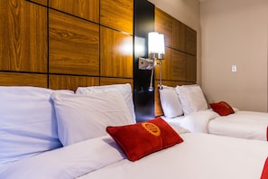 Habitación de hotel con dos camas, ropa de cama blanca, una almohada roja y una lámpara fijada a la pared.