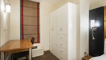 A hotel room with a wooden desk, a white wardrobe, a black wardrobe, a bed, and a wall-mounted light fixture.