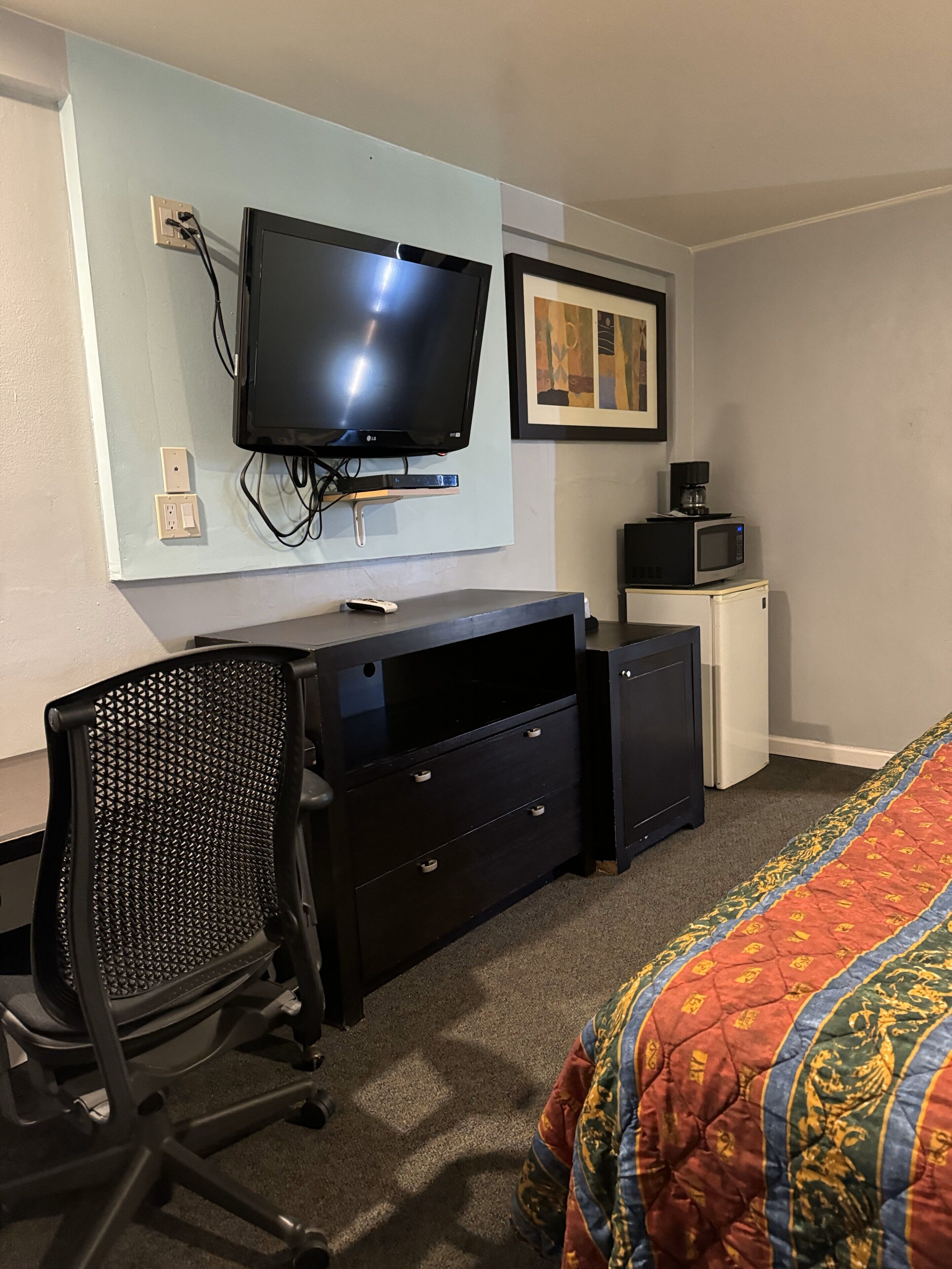 Habitación de hotel con televisión de pantalla plana en la pared, un escritorio con cajones, una silla, un microondas y una cama con una manta colorida.