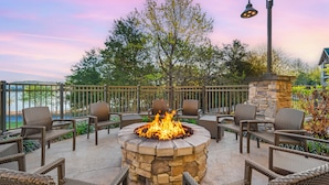 An outdoor patio with a stone fire pit surrounded by wicker chairs, overlooking a body of water.