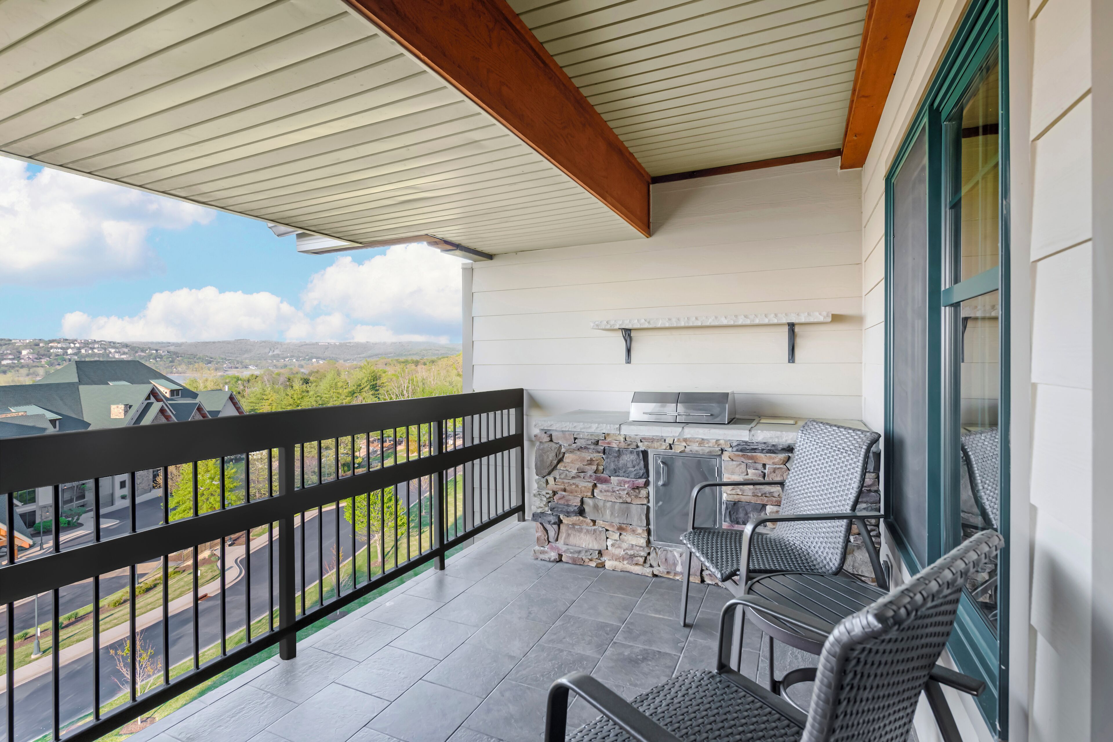 A covered balcony with a stone wall, a grill, and two wicker chairs.