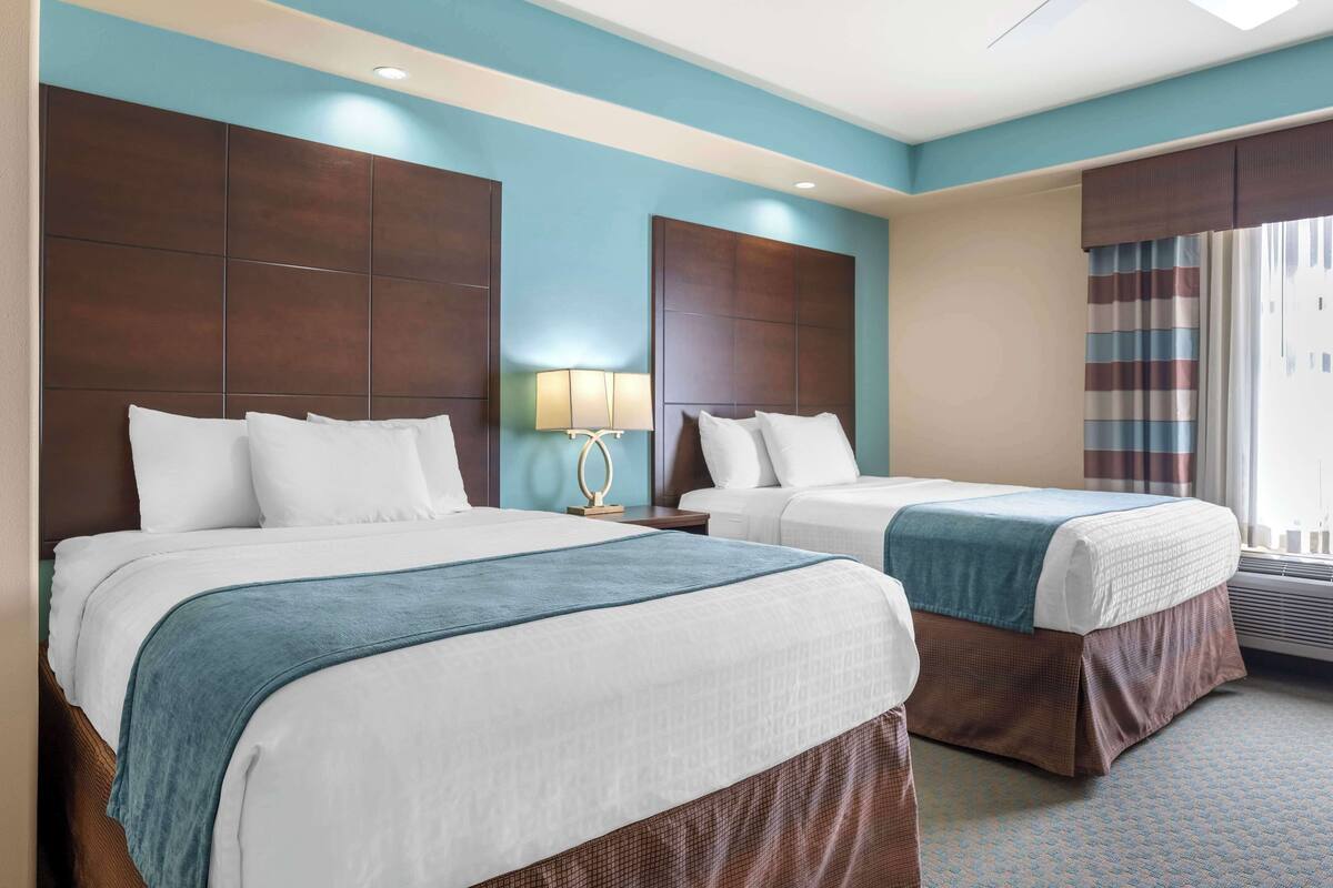 Una habitación de hotel con dos camas, un ventanal con cortinas, un ventilador de techo y una lámpara de pared.
