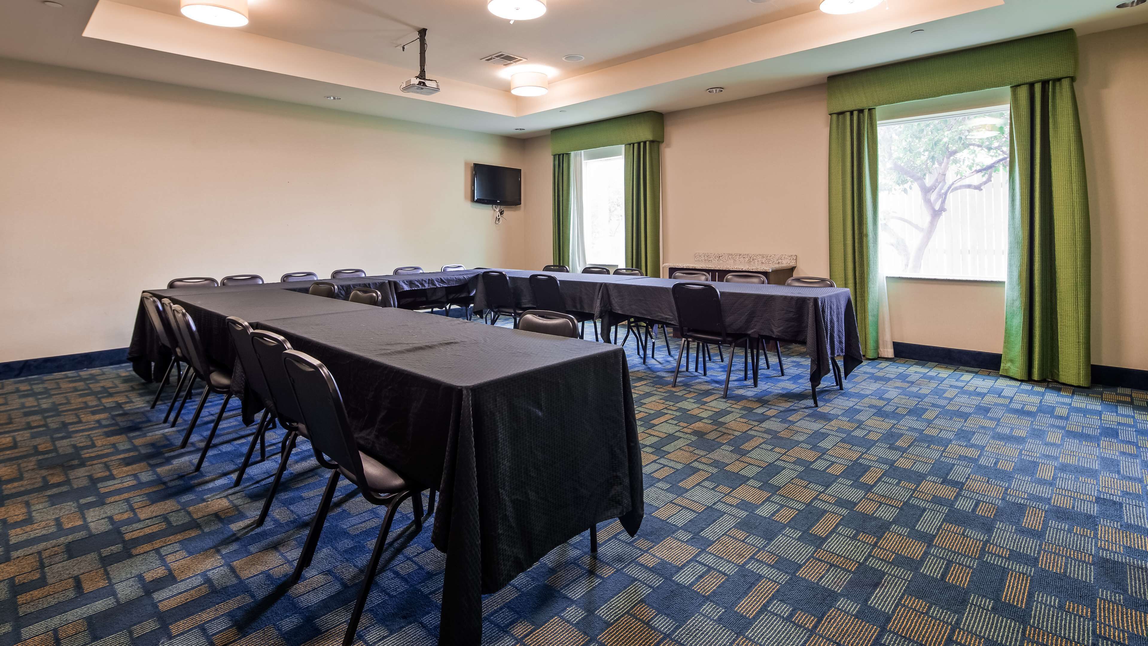 A conference room with multiple tables and chairs, a flat-screen TV, and large windows with green curtains.