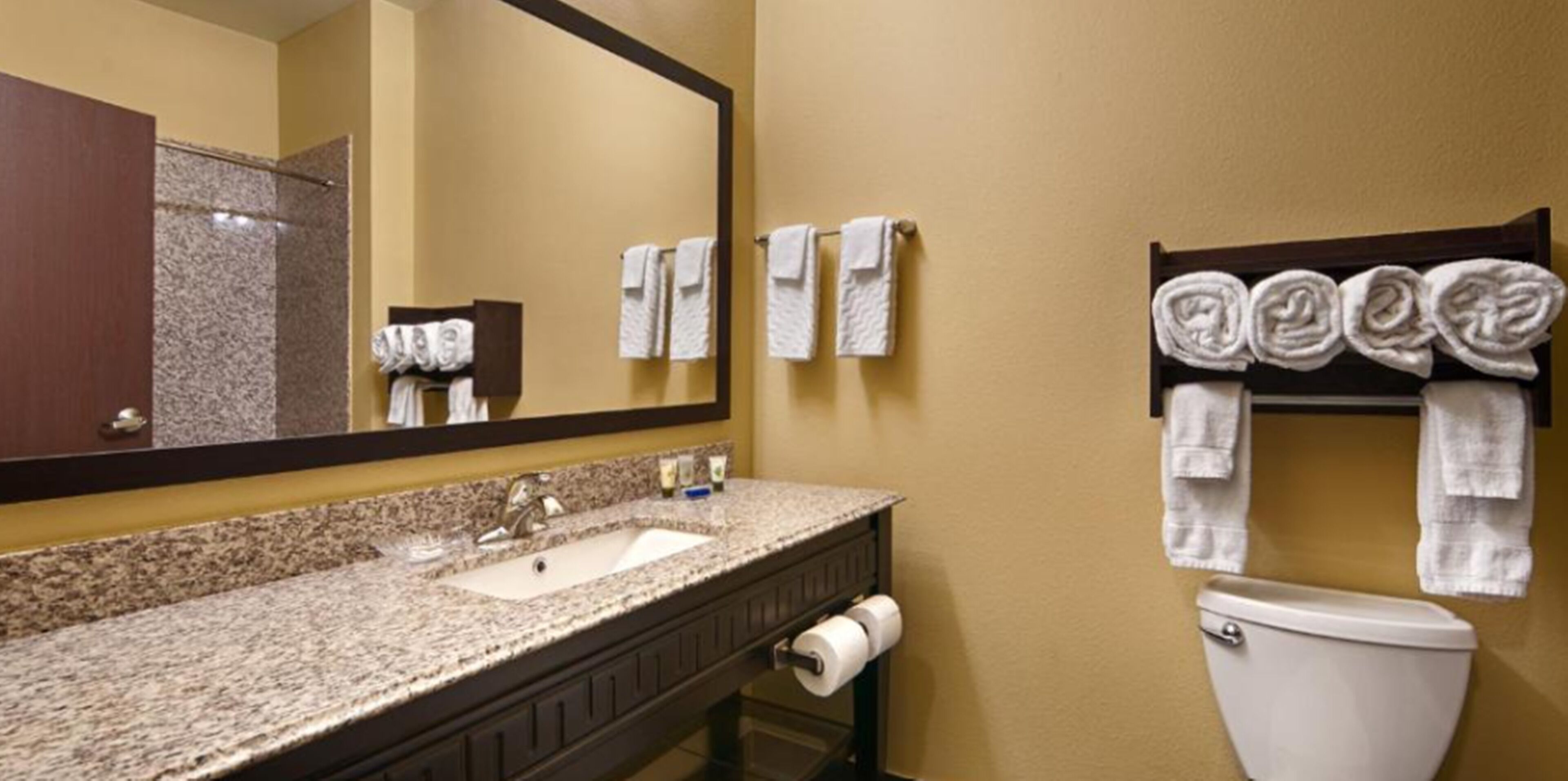 Un baño de hotel con lavamanos de encimera de granito, un espejo, un inodoro y toallas colgadas en la pared.