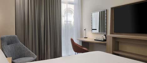 A hotel room with a large bed, a desk with a flat-screen TV, a chair, and a large window with curtains.