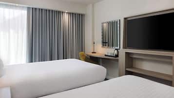 A hotel room with two beds, a desk, a chair, a TV, and a large window with curtains.