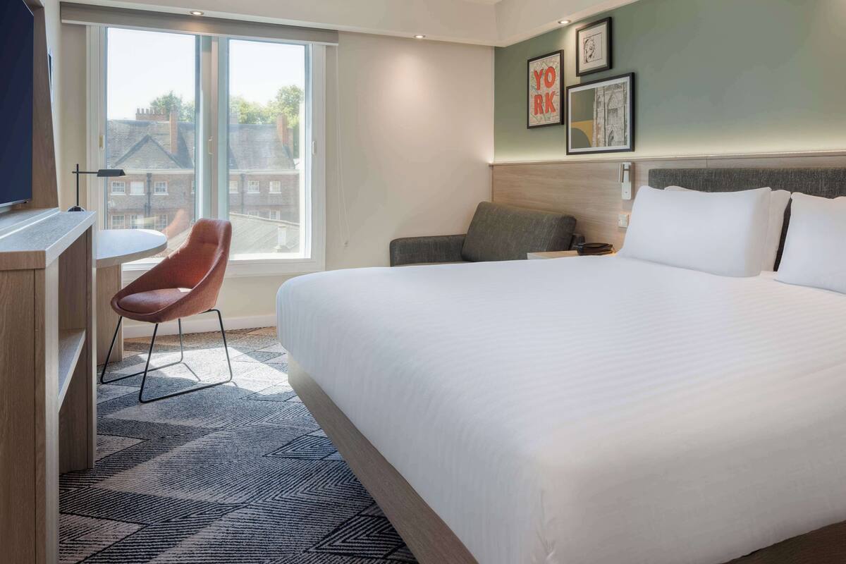 A hotel room with a large bed, a desk with a chair, a sofa, and a large window offering a view of buildings outside.